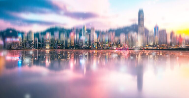 A blurry cityscape with reflections in the water. The sky is a soft pink and blue with clouds.