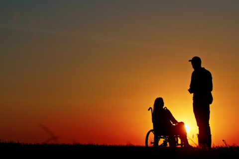 Silhouette of a person standing beside a woman in a wheelchair during a beautiful sunset. A scene symbolizing support, companionship, and hope under the golden glow of dusk.