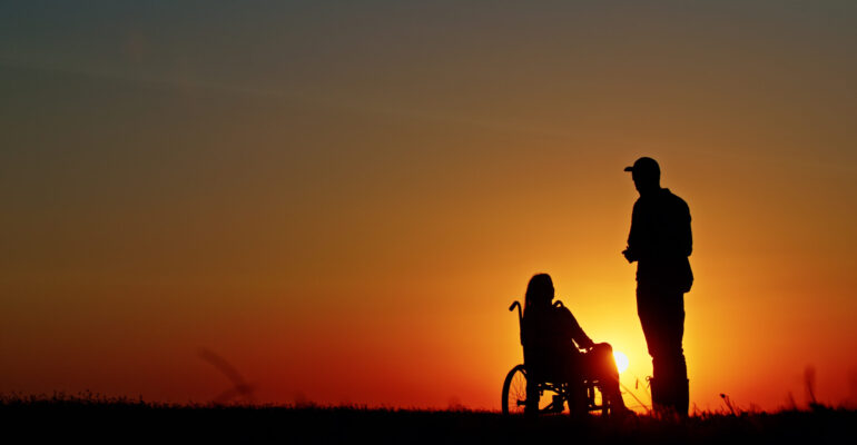 Silhouette of a person standing beside a woman in a wheelchair during a beautiful sunset. A scene symbolizing support, companionship, and hope under the golden glow of dusk.